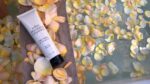 Tube of John Masters nærende hårmaske med Rose & Abrikos 148 ml placeret på en træplade omgivet af gule og hvide rosenblade, der flyder i vand.