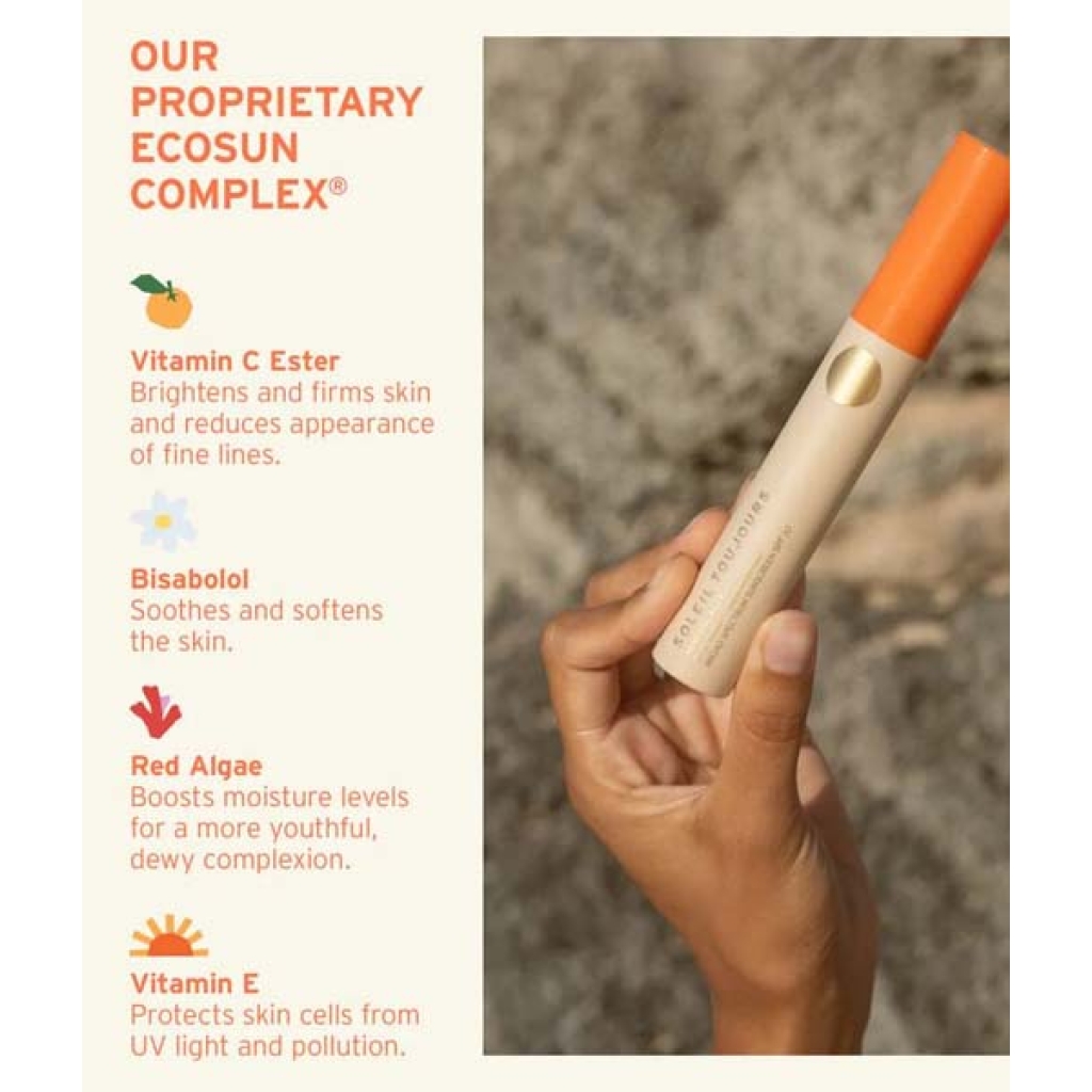 En hånd holder et Soleil Toujours Mineral Ally Eye Glow + Illuminator SPF 30 ml produkt med en orange hætte. Teksten til venstre viser ingredienser: C-vitaminester, Bisabolol, Rødalger og E-vitamin, der fremhæver deres fordele for hudpleje.