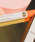 Nærbillede af Soleil Toujours Mineral Ally Eye Glow + Illuminator SPF 30 ml, præsenteret på en reflekterende overflade ved siden af en pink genstand.