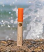 En tube solcreme med en orange hætte mærket "Soleil Toujours Mineral Ally Eye Glow + Illuminator SPF 30 ml" står oprejst på en stenet strand med havbølger, der plasker i baggrunden.