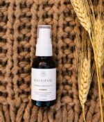 En lille flaske Innersense Harmonic Healing & Treatment Oil Skin, Scalp & Hair 25ml sidder på en tekstureret brun overflade, ledsaget af hvedestilke.