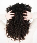 Person med langt, krøllet mørkt hår set fra ryggen, hænderne placeret i håret mod en hvid baggrund, der viser den voluminøse effekt af Innersense I Create Volume - Volumizing Lotion 295ml.