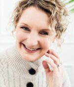 En person med krøllet hår og et varmt smil, sandsynligvis forstærket af Innersense I Create Volume - Volumizing Lotion 295ml, er iført en striktrøje med store knapper. De rører blidt ved deres ansigt med den ene hånd.