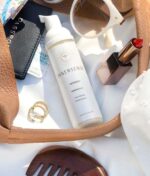 Forskellige ting, herunder en flaske Innersense Refresh Dry Shampoo - Skum til pulver 70 ml, læbestift, solbriller, ringe, et pas og andre vigtige rejseartikler er arrangeret på en overflade.