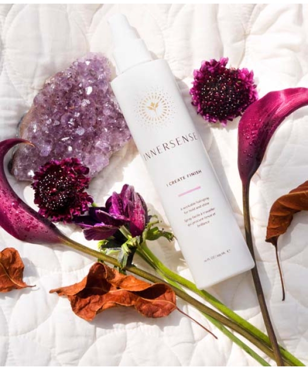 En hvid flaske Innersense I Create Finish - Shine Spray 295ml er placeret mellem lilla blomster, en lilla krystalklase og brune tørrede blade på et hvidt quiltet stof.