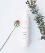 En hvid flaske Innersense I Create Hold - Styling gel 295ml placeres ved siden af en lille lyserød blomst og noget grønt løv på en hvid baggrund.