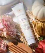 En tube med Innersense Hydrating Hair Mask - Repair & Revitalize 177ml er placeret på et tekstureret stof, omgivet af en pink rose, et cremefarvet græskar og en brun trækam.