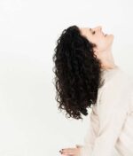 En person med krøllet mørkt hår vipper hovedet tilbage og ser opad, iført en lys langærmet skjorte. Deres lokker fremstår frisk rensede, måske takket være Innersense Color Awakening Hairbath 295ml (sulfatfri), sat mod en almindelig hvid baggrund.