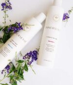 To Innersense hårproduktflasker, en til "Quiet Calm Curl Control" og en til "Innersense I Create Volume - Volumizing Lotion 295ml," vises på en hvid overflade prydet med små lilla blomster og grønt løv.