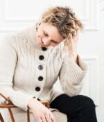 En person med krøllet hår, forstærket af Innersense Inner Peace Whipped Creme Texturizer - Styling creme 96g, sidder og smiler med hovedet vippet ned og hånden rører ved håret, iført en beige sweater med store knapper.