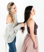 To kvinder står. En af dem, iført en grå sweater og jeans, er ved at flette det lange mørke hår på den anden kvinde, som er iført en brun tank top og en lyserød nederdel. Begge smiler, deres lokker er frisk revitaliseret med Innersense Refresh Dry Shampoo - Skum til pulver 70 ml.
