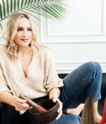 En kvinde med lyst hår, iført en beige sweater og blå jeans, sidder på en sofa med en skål og en støder, med Innersense Refresh Dry Shampoo - Skum til pulver 70 ml og en grøn plante i baggrunden.