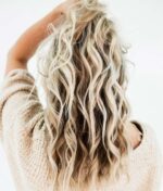 En person med bølget, blond hår ses bagfra, iført en lys striktrøje og løfter den ene hånd for at røre ved deres hår, som gløder med sundheden og livligheden fra Innersense Color Awakening Hairbath 295ml (sulfatfri), sat mod en bleg baggrund.