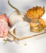 En bakke viser en krukke med Innersense Inner Peace Whipped Creme Texturizer - Styling creme 96g, et åbent låg, en lille børste, et hvidt græskar, lyserød blomst og en vase med en orange krysantemum.