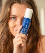 En kvinde med fregner holder en blå flaske COOLA Full Spectrum 360° Refreshing Water Mist SPF 15 50ml op i nærheden af hendes ansigt, hvilket delvist skjuler det ene øje.