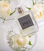 En flaske "Ærlig P03 - Eau de Parfum 100 ml glasflaske" ligger blandt hvide roser og babypusteblomster i klare glasskåle.