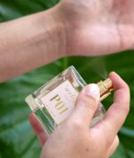 En person, der sprøjter en klar flaske parfume mærket "Ærlig P01 - Eau de Parfum 100 ml glasflaske" på sit håndled mod en baggrund af grønne blade.