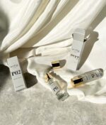 To parfumeflasker, mærket "P0" og "Ærlig P02 - Eau de Parfum 100 ml glasflaske", er udstillet på et blødt hvidt stof med deres respektive æsker i nærheden. Den ene flaske er åben og afslører en guldsprøjtedyse.