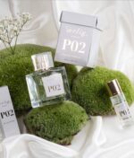 En parfumeflaske mærket "Ærlig P02 - Eau de Parfum 100 ml glasflaske" placeret på grønt mos, ledsaget af et lille hætteglas, en æske og en blomsterkvist, sat mod en hvid stofbaggrund.