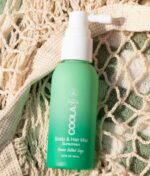 En grøn flaske COOLA Scalp & Hair Mist Sunscreen SPF30 (vandafvisende 80 min) 60 ml, mærket "Ocean Salted Sage," hviler på en beige tekstur klud.