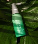 Grøn flaske COOLA Scalp & Hair Mist Sunscreen SPF30 (vandafvisende 80min) 60ml med Ocean Salted Sage-duft, placeret på en stor, våd grøn bladbaggrund.