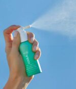 Håndsprøjtning COOLA Scalp & Hair Mist Sunscreen SPF30 (vandafvisende 80min) 60ml mod en klar blå himmel.