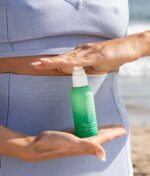 En person i et blåt outfit holder en grøn flaske COOLA Scalp & Hair Mist Sunscreen SPF30 (vandafvisende 80min) 60ml, med den ene hånd over flasken og den anden hånd under den, nær en strand.