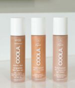 Tre flasker COOLA Mineral Rōsilliance® Tinted Moisturizer solcreme - Golden hour SPF 30 44ml står oprejst på en hvid overflade, der hver specificerer forskellige nuancer og funktioner, hvilket indikerer, at de er økologiske hudplejeprodukter med solcreme.