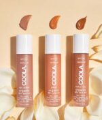 Tre flasker COOLA Mineral Rōsilliance® Tinted Moisturizer solcreme - Golden hour SPF 30 44ml i lyse/medium, gyldne og medium/dybe nuancer, omgivet af rosenblade, med farveprøver af hver nuance over den tilsvarende flaske.