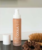 En flaske COOLA Mineral Rōsilliance® Tinted Moisturizer solcreme - Golden hour SPF 30 44ml står på en badeværelsesskranke ved siden af en brun hårscrunchie og en træhårbørste.