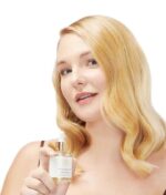 En person med langt lyst hår holder en dråbeflaske med Innersense I Create Shine 50 ml, smilende let og kigger på kameraet.