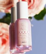 En lyserød flaske COOLA Dew Good Illuminating Serum Sunscreen med Probiotic SPF 30, mærket "COOLA Dew Good Illuminating Serum Sunscreen with Probiotic SPF 30," afspejles på baggrund af lyserøde roser, der ikke er i fokus.