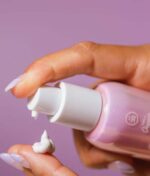 Håndpresser COOLA Dew Good Illuminating Serum Sunscreen med Probiotic SPF 30 fra en pumpeflaske på en finger mod en lilla baggrund.