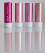 Fire COOLA Mineral Liplux® SPF30 - Nude Beach er linet op, hver med et andet mærke: "Firecracker", "Nude Beach", "Summer Crush" og "Skinny Dip." Læbepomaderne er lyserøde og metalliske med hvide bunde.