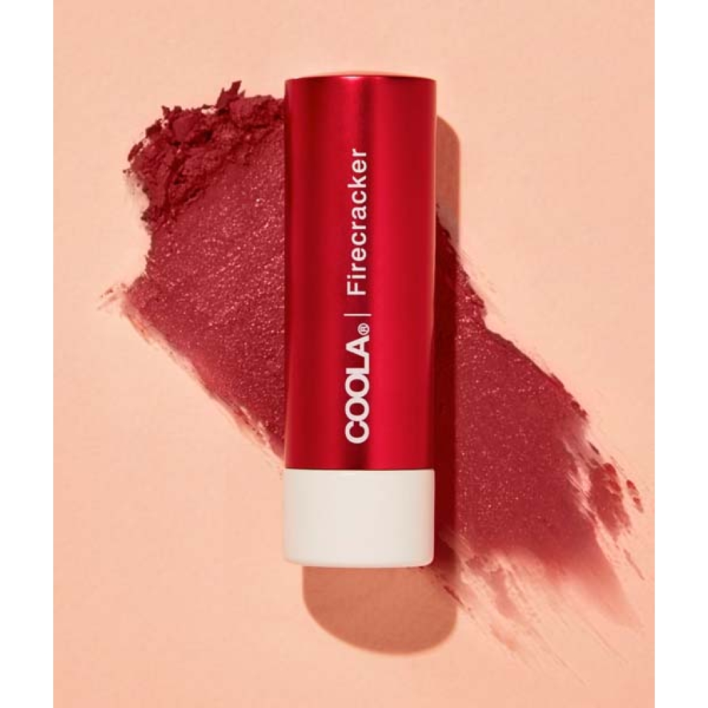 Et rødt rør med COOLA Mineral Liplux® SPF30 - Firecracker placeres diagonalt på en lys ferskenbaggrund med en udtværing af produktet ved siden af.