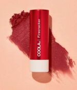 Et rødt rør med COOLA Mineral Liplux® SPF30 - Firecracker placeres diagonalt på en lys ferskenbaggrund med en udtværing af produktet ved siden af.