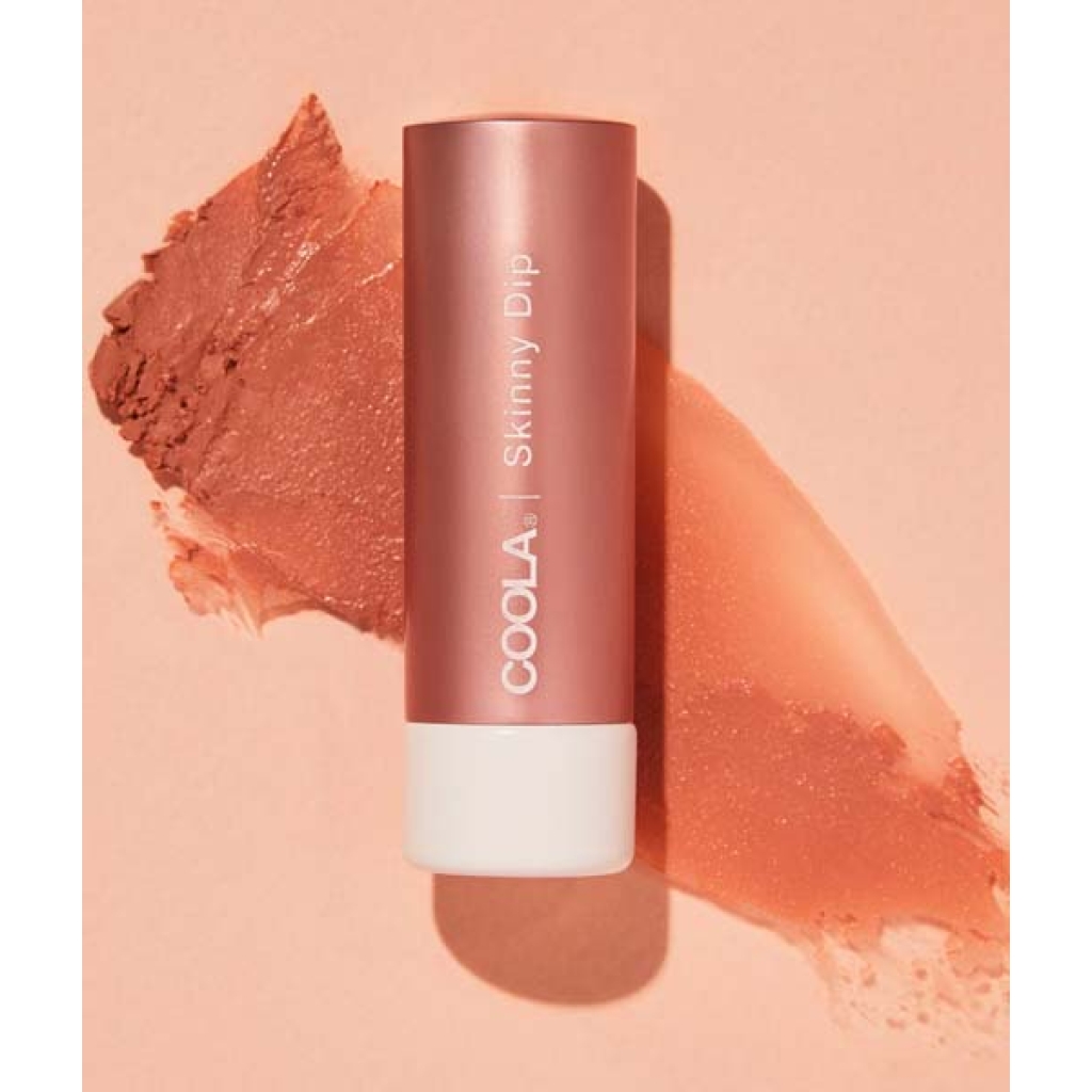En tube COOLA Mineral Liplux® Tinted Lip Balm SPF 30 - Skinny Dip liggende på en ferskenbaggrund med et stryg af balsam ved siden af.