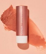 En tube COOLA Mineral Liplux® Tinted Lip Balm SPF 30 - Skinny Dip liggende på en ferskenbaggrund med et stryg af balsam ved siden af.