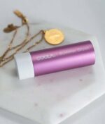 Et lyserødt rør af COOLA Mineral Liplux® SPF30 - Nude Beach er placeret på en hvid marmoroverflade ved siden af to guldhalskæder.