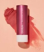En pink COOLA Mineral Liplux® SPF30 - Summer Crush tube ligger på en lys pink overflade med en farveprøve af læbepomadefarven smurt ud ved siden af.