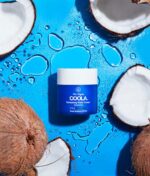 En blå krukke med COOLA Refreshing Water Cream SPF 50 - 44ml sidder på en levende blå overflade, prydet med friske kokosnøddehalvdele og legende stænk af vand.