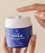 En hånd rører en lille mængde COOLA Refreshing Water Cream SPF 50 - 44ml på dispenserlåget på en blå beholder. Produktet har SPF 50, er 70%+ økologisk og tilbyder bredspektret beskyttelse.