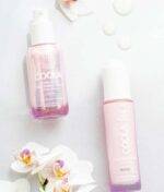 To COOLA hudplejeprodukter er elegant placeret på en hvid overflade prydet med orkidéblomster. Fremhævet er COOLA Dew Good Illuminating Serum Sunscreen med Probiotic SPF 30 og Sun Silk Drops solcremen, som begge tilbyder solbeskyttelse. Navnlig COOLA Dew Good Illuminating Serum Sunscreen med Probiotic SPF 30 giver avancerede hudplejefordele.