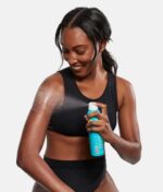 En kvinde i sort sports-bh og -underdel påfører COOLA Classic Sunscreen Spray Parfume-Free SPF 50 177 ml på sin arm.