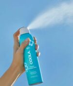 Hånd, der holder en COOLA Classic Sunscreen Spray Duftfri SPF 50 177 ml flaske, sprøjter produktet mod en blå himmel.
