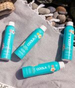 Fire blå flasker COOLA Classic Sunscreen Spray Parfume-Free SPF 50 177 ml placeres på et stribet klæde med sten i baggrunden.