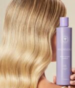 En person med bølget blond hår holder en 946 ml lilla flaske Innersense Bright Balance Hairbath, en shampoo.