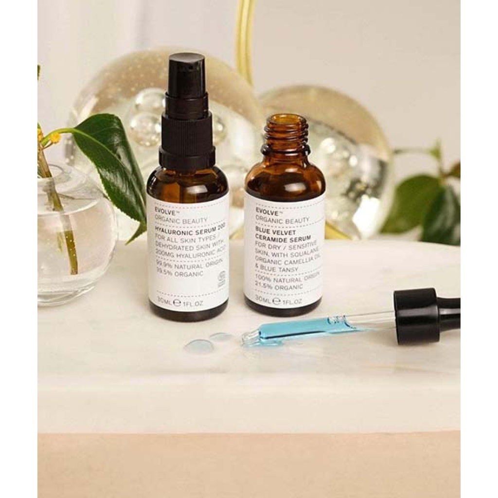 To små flasker fra Evolve Hydrate & Soothe Bundle på en badeværelsesdisk med en dråbe og botaniske dekorationer i baggrunden.
