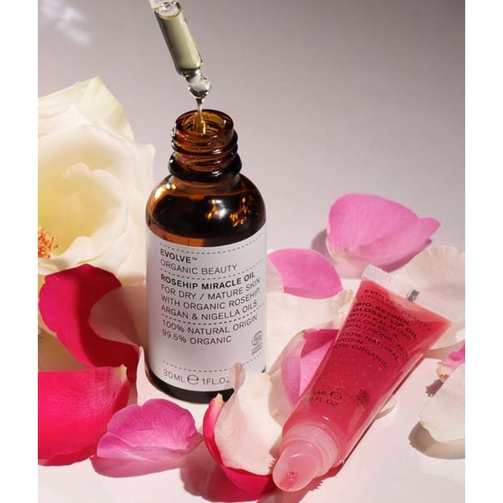 Dropper flaske Evolve Miracle Face & Lip Oil Duo med etiket, omgivet af spredte rosenblade og en hvid rose, på en lys baggrund.