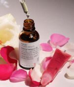 Dropper flaske Evolve Miracle Face & Lip Oil Duo med etiket, omgivet af spredte rosenblade og en hvid rose, på en lys baggrund.